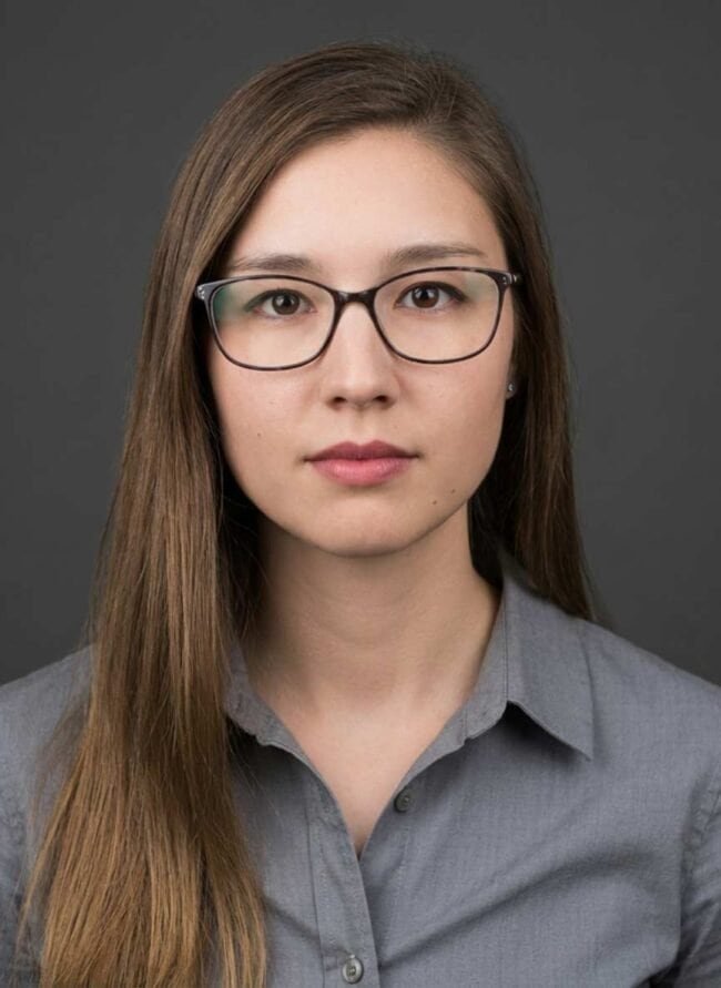 High-quality professional portrait of a young woman with glasses, grey shirt, and a neutral background.